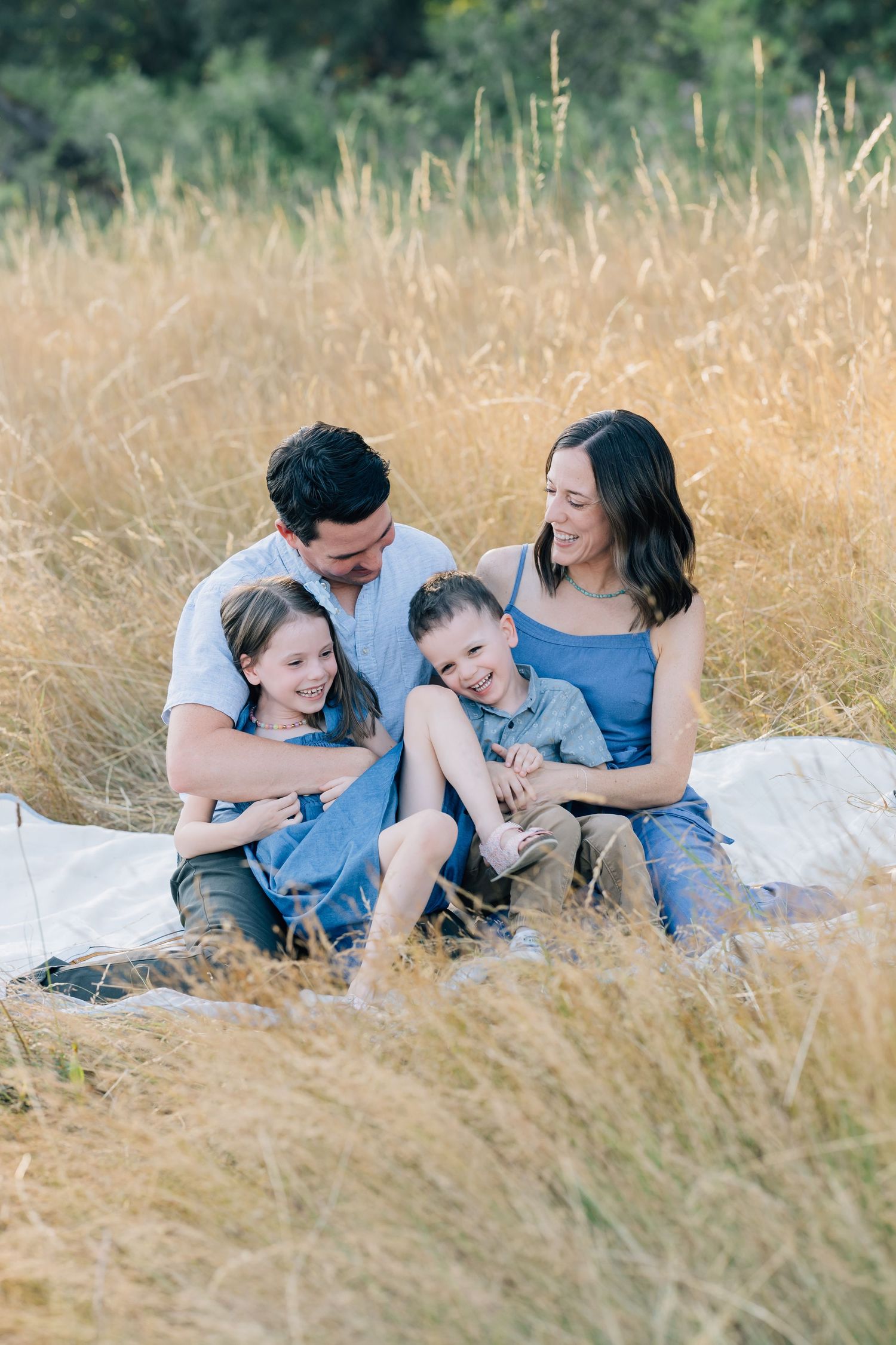 A happy family of four sits together in golden tall grass, smiling and embracing during an outdoor photo session.