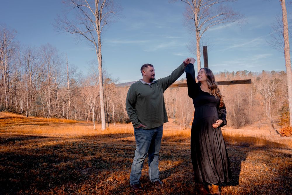 Couple dancing outdoors during maternity photo shoot in autumn forest setting with blue sky.