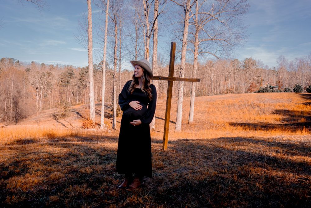 Pregnant woman in black dress and cowboy hat standing beside wooden cross in autumn field.