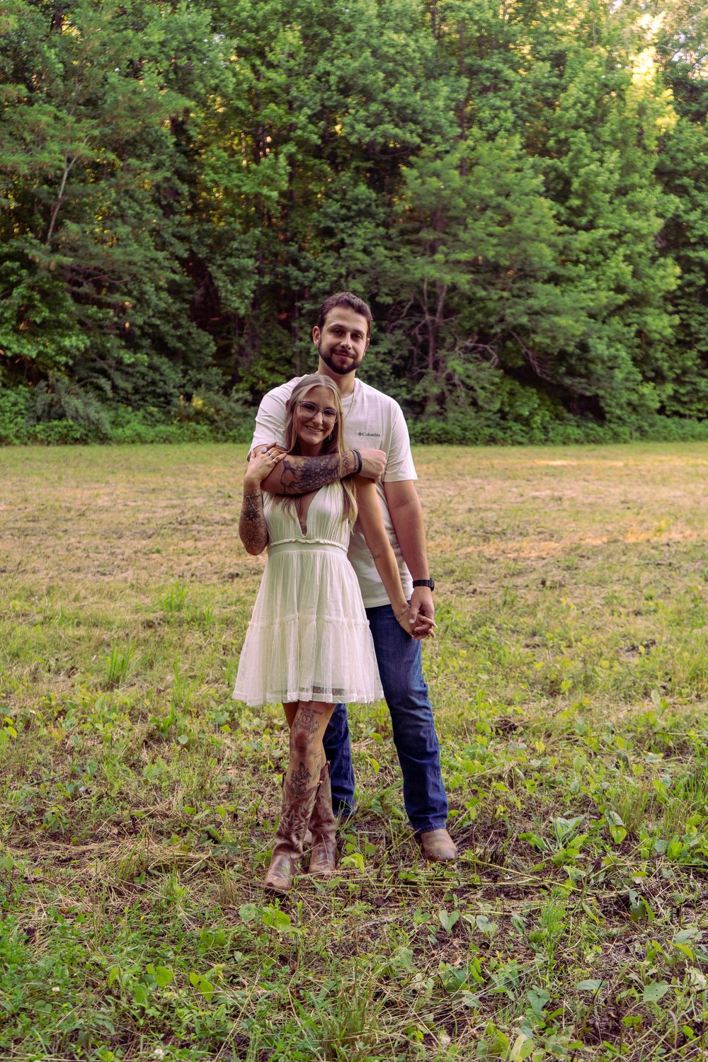 Happy couple embracing in green meadow surrounded by lush forest trees at sunset.