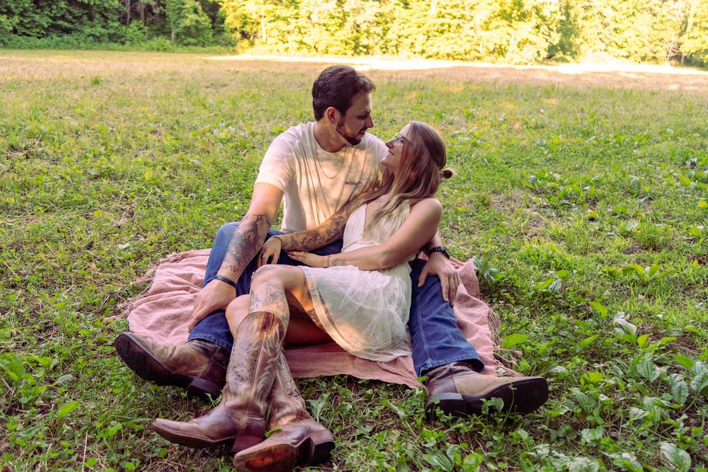 Couple sitting on blanket in green park, sharing romantic moment in cowboy boots and casual summer outfits.