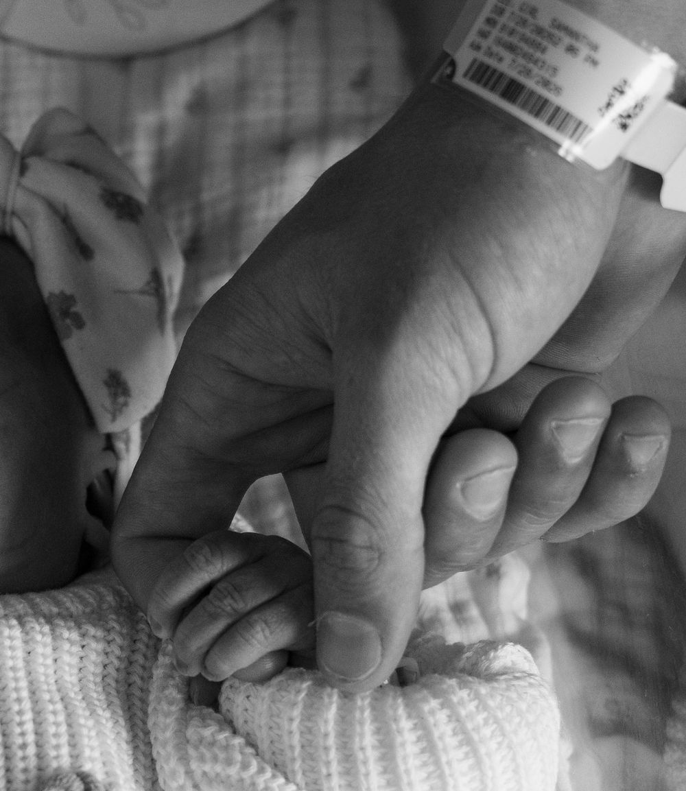 Black and white photo of a newborn baby grasping an adult's finger, with a hospital ID bracelet visible.