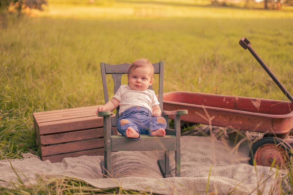 Adorable baby sitting in small wooden chair in golden field next to red wagon outdoors.