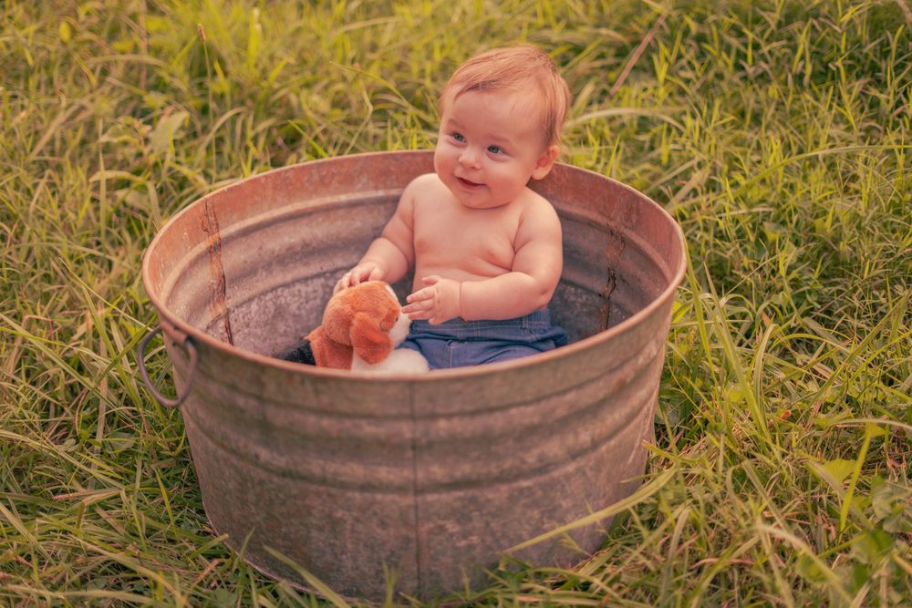 A smiling baby sits in a rustic metal tub outdoors in green grass, holding a toy.