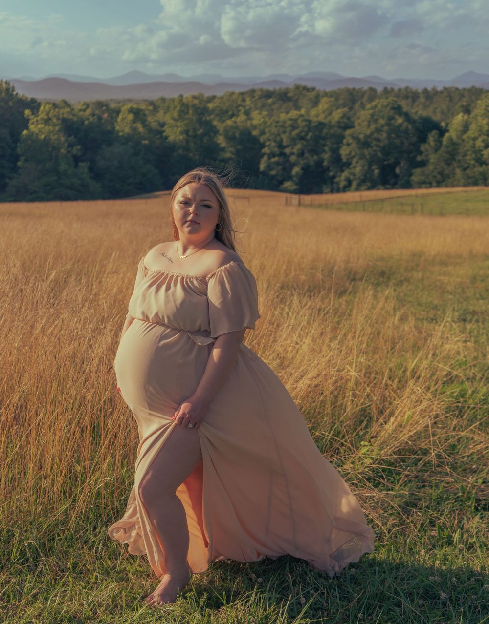 Pregnant woman in flowing peach maternity gown posing in golden field with mountain backdrop.