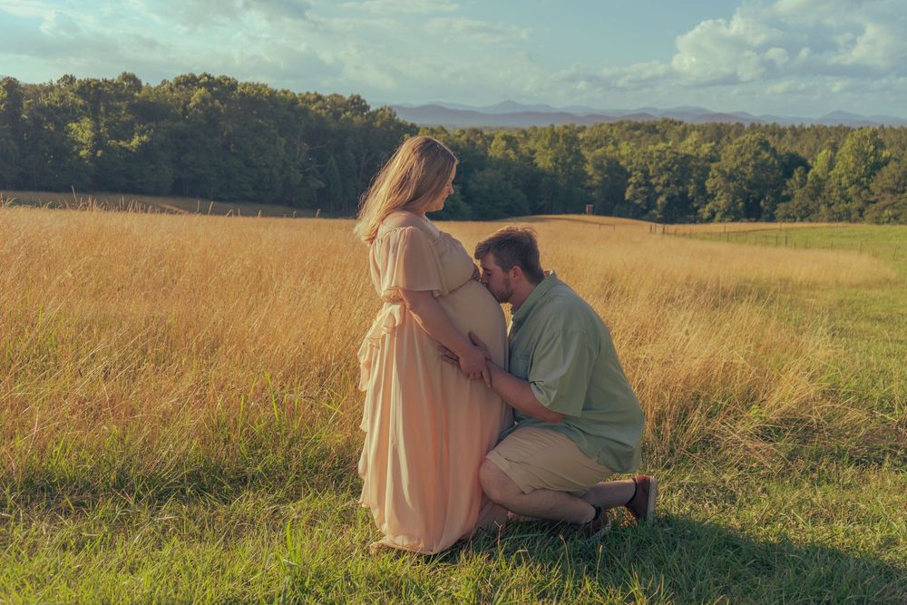 Man kneeling and kissing pregnant partners belly in scenic golden field at sunset.