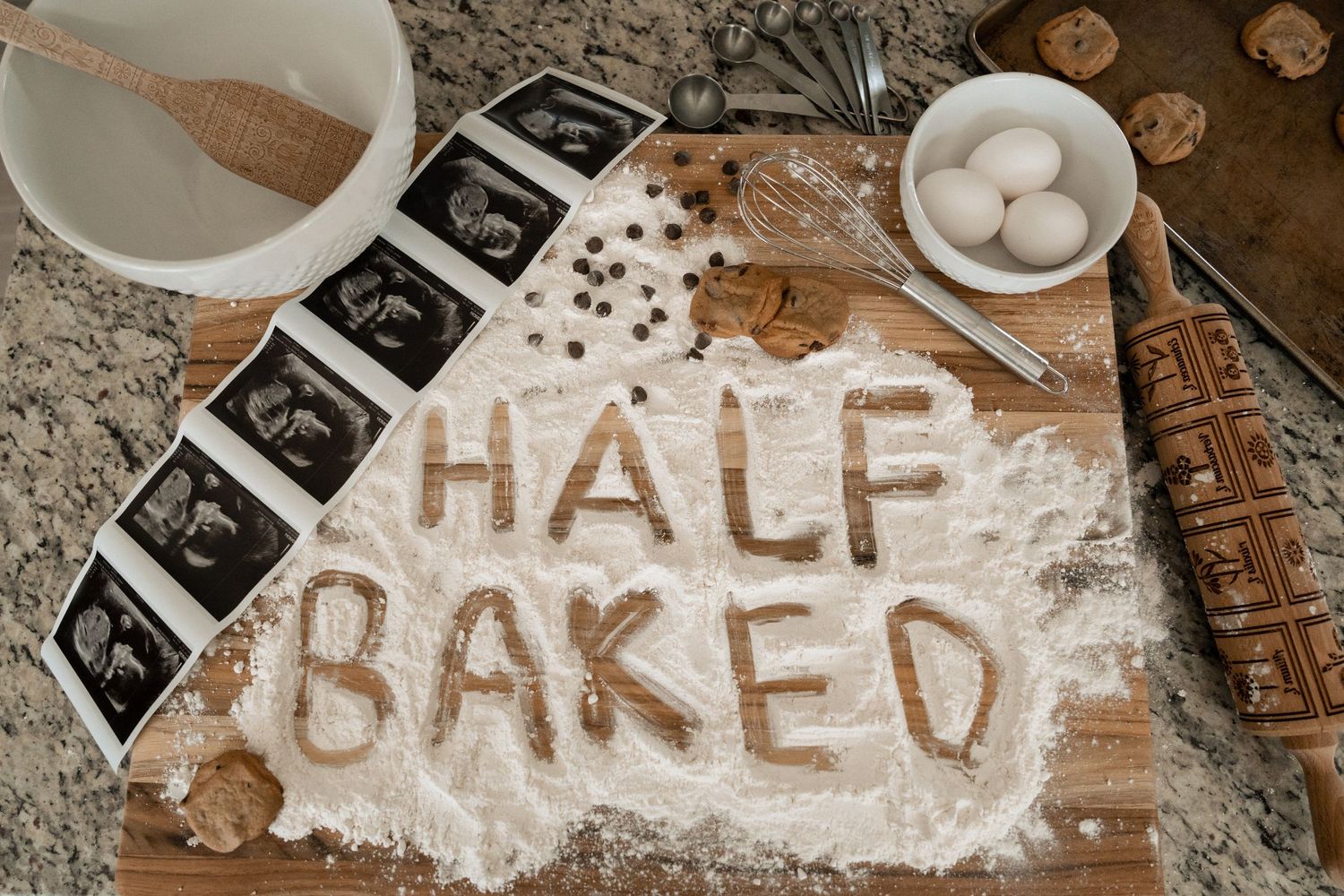 Flour spelling HALF BAKED on cutting board with ultrasound photos, eggs, chocolate chips, pregnancy announcement.