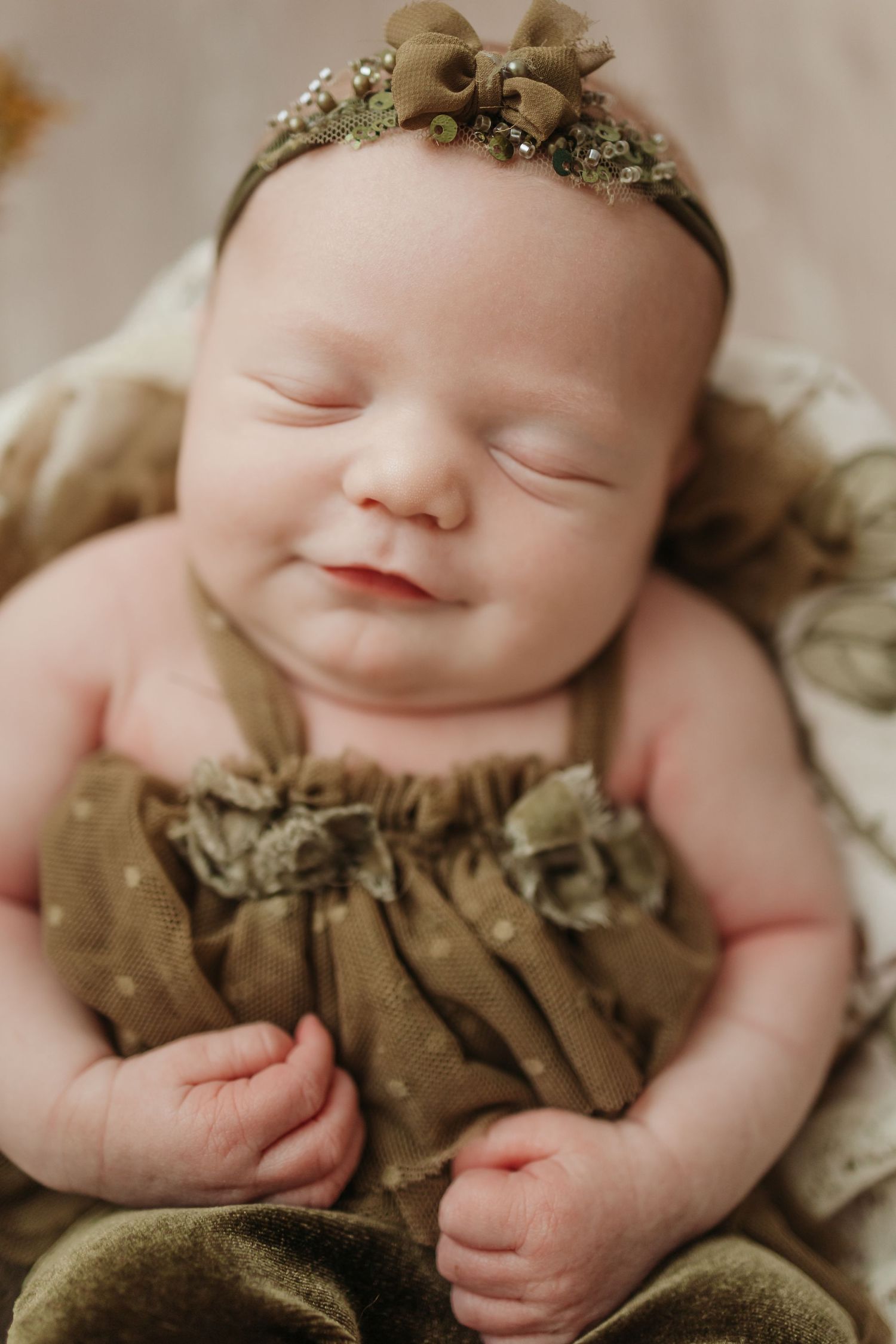 Smiling newborn baby girl wearing olive green ruffled outfit and floral headband, eyes closed, posed in studio portrait.