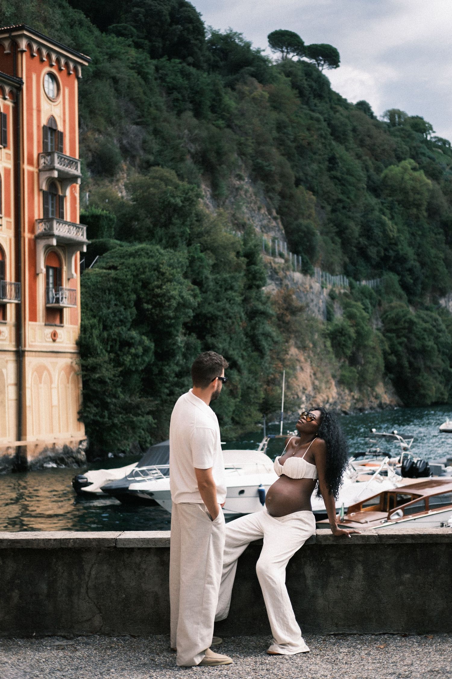Couple standing on a lush green balcony overlooking a scenic lake and mountain landscape in Italy.
