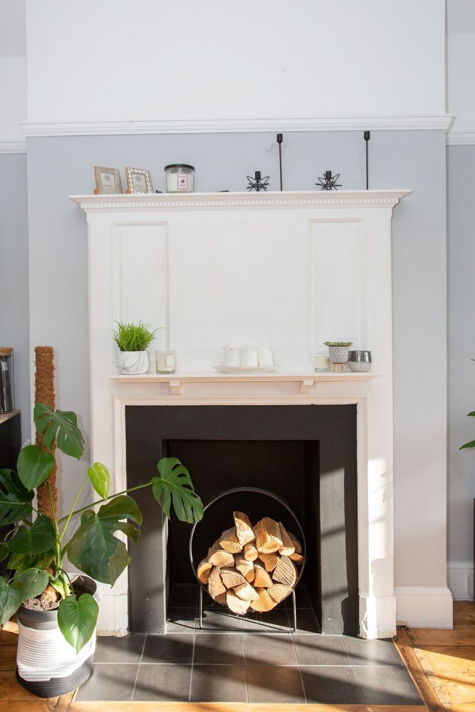 White fireplace mantel with decorative items and stacked firewood in sunlit living room.
