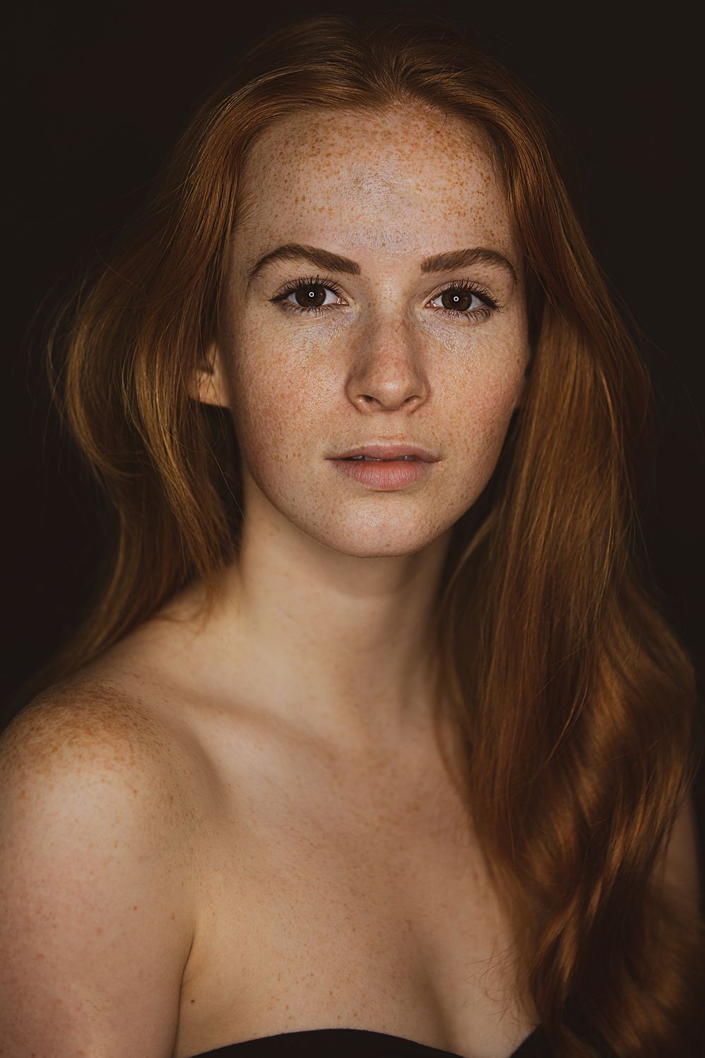Portrait of young woman with long red hair and freckles against dark background.