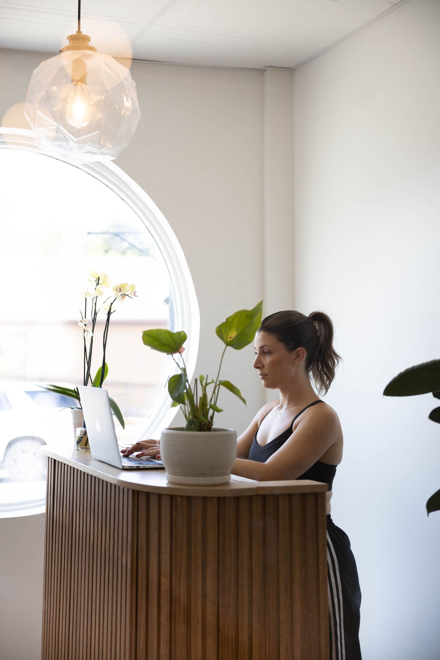 Woman in athletic wear working on laptop at wooden reception desk with plants and round window in bright modern studio.