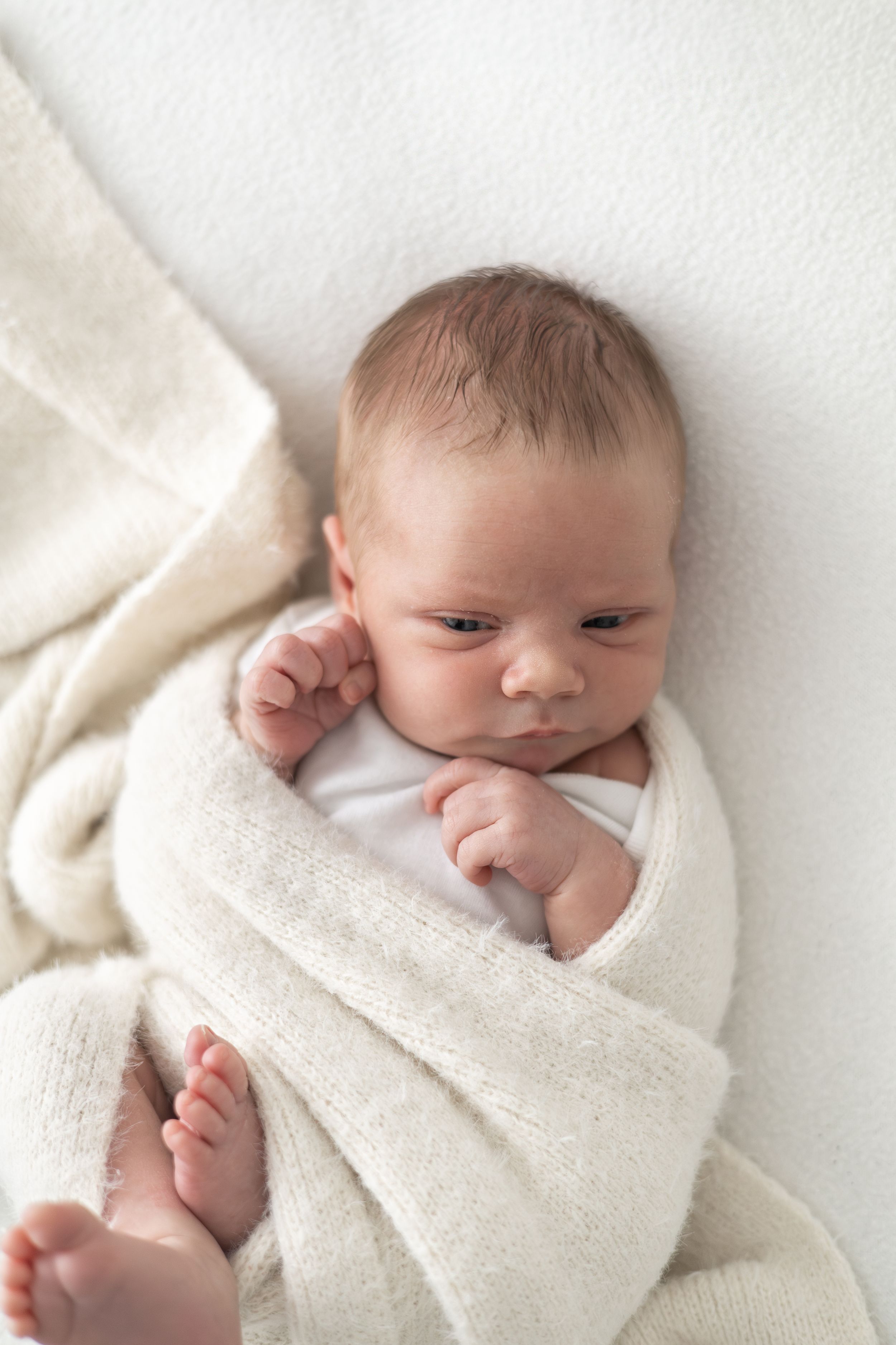 Newborn baby swaddled in a soft cream blanket, lying on a white surface with tiny feet peeking out.