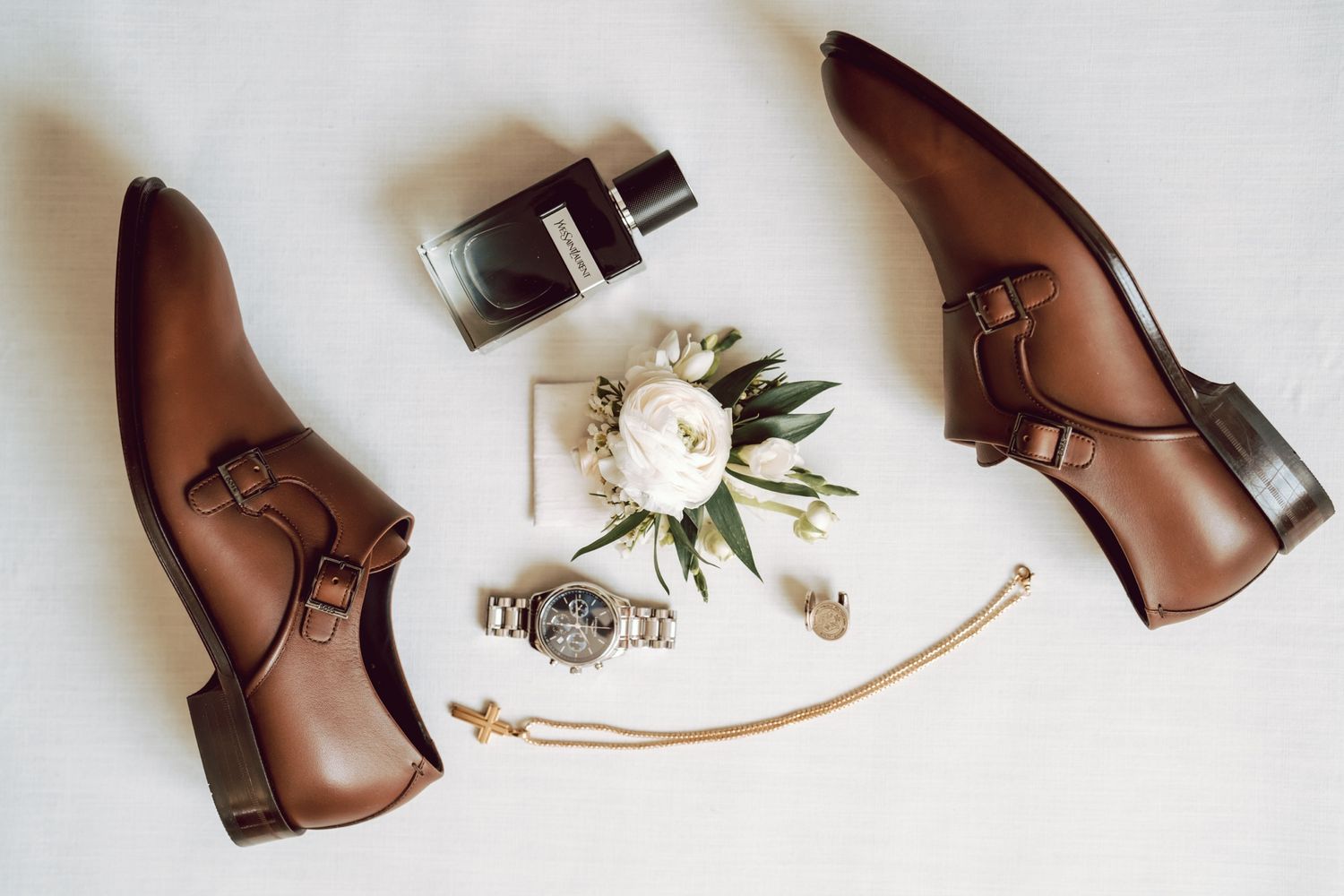 Brown leather monk strap dress shoes with a boutonniere, cologne, silver watch, and accessories arranged on white background.