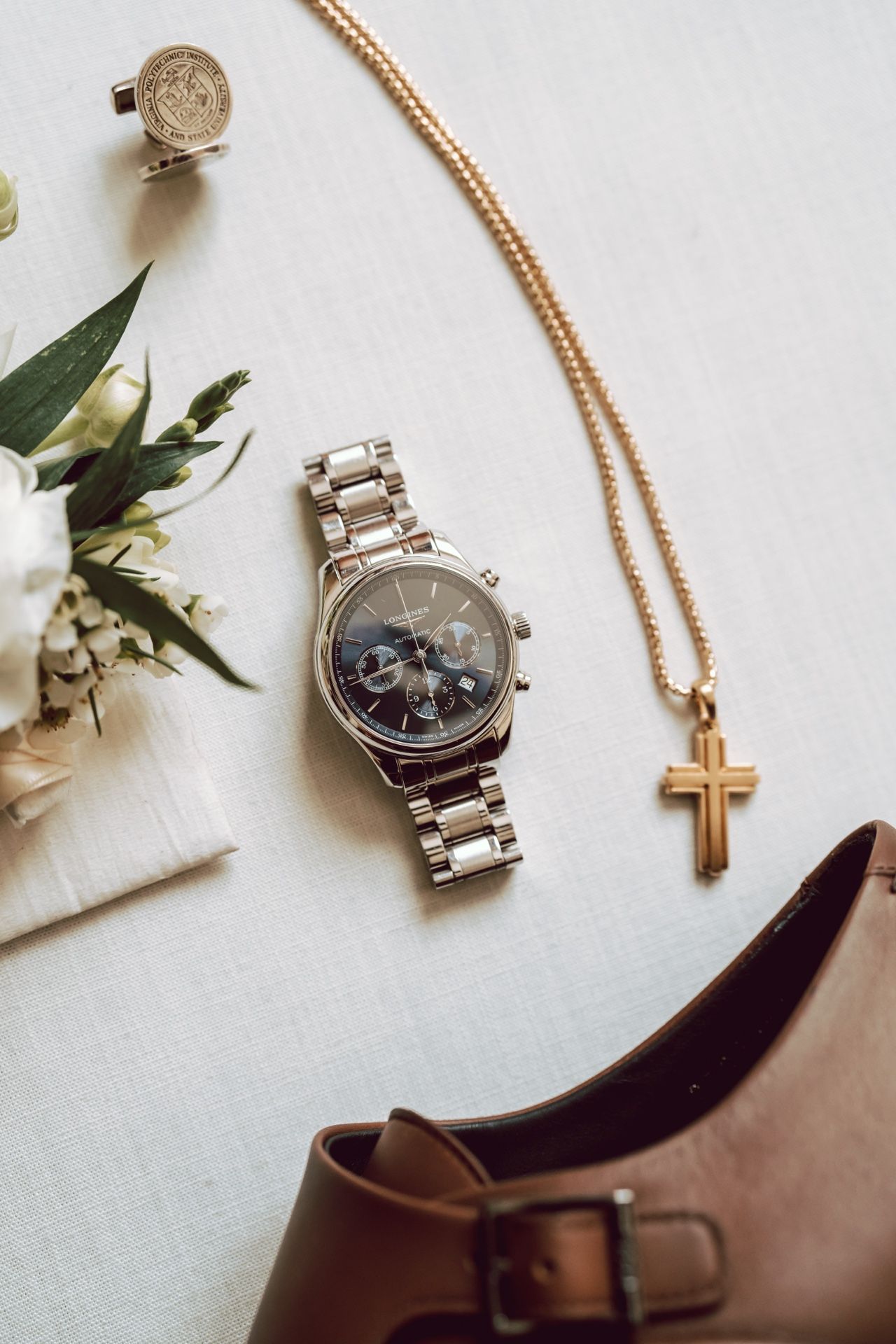 Groom's wedding accessories flat lay with silver watch, gold cross necklace, cufflinks, boutonniere, and brown leather shoes.
