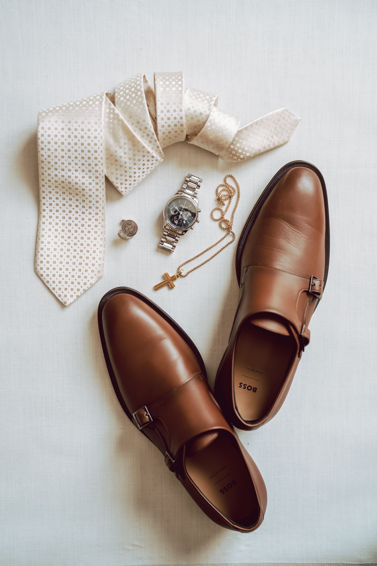 Brown leather monk strap dress shoes beside a polka dot tie, silver watch, gold cross necklace, and cufflinks on white surface.