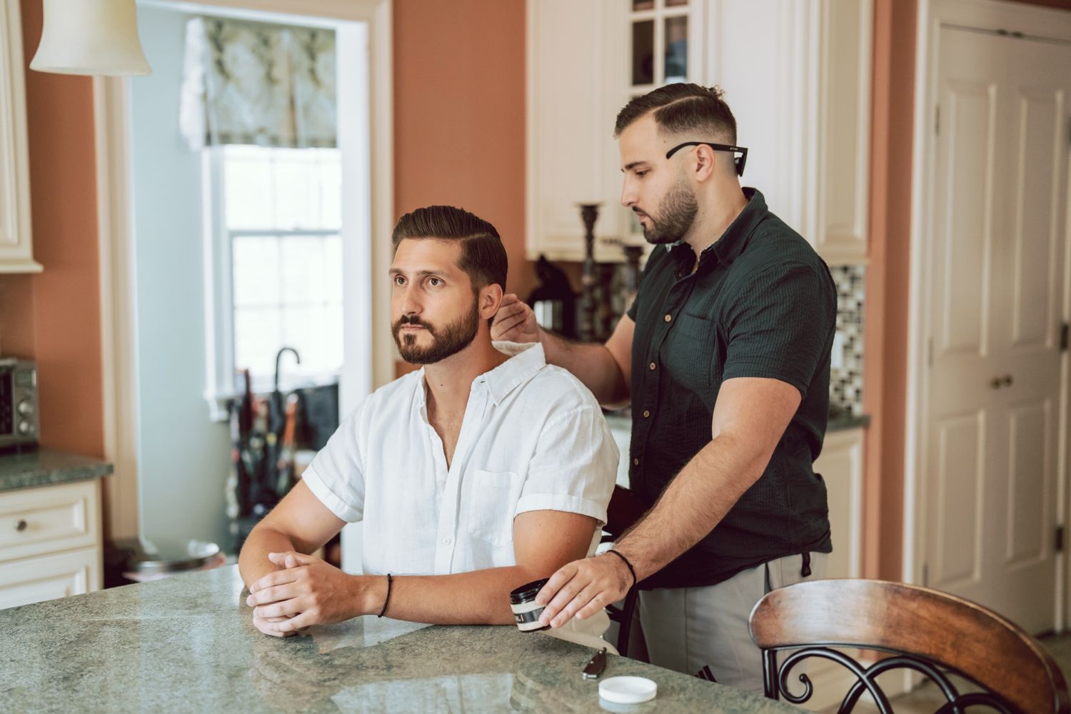 Two men in a kitchen, one in a white shirt sitting at a granite counter while another in black stands behind him.