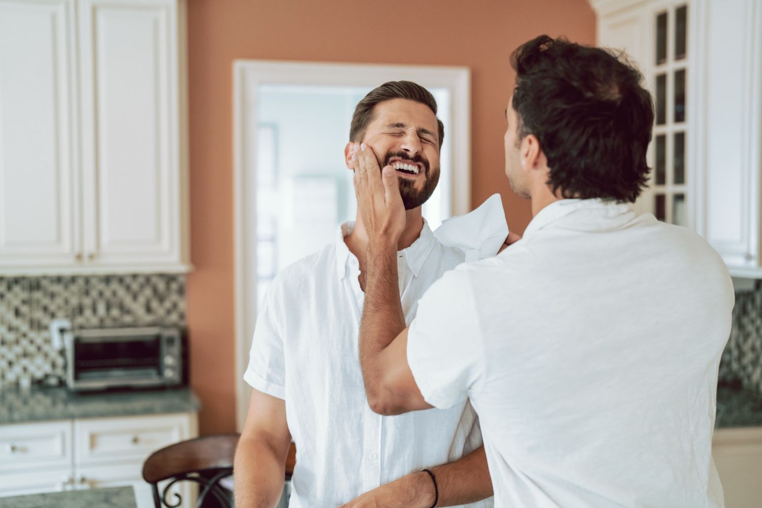 Two men sharing a tender moment in a bright kitchen, one gently touching the other's face while smiling.