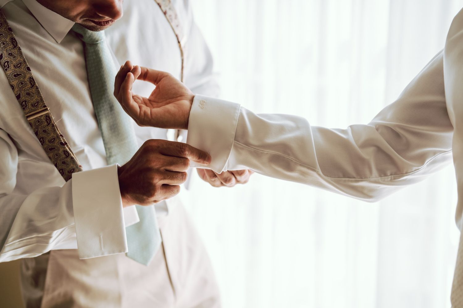 Two men in white dress shirts and suspenders, one adjusting the other's cufflinks, with a green tie visible.