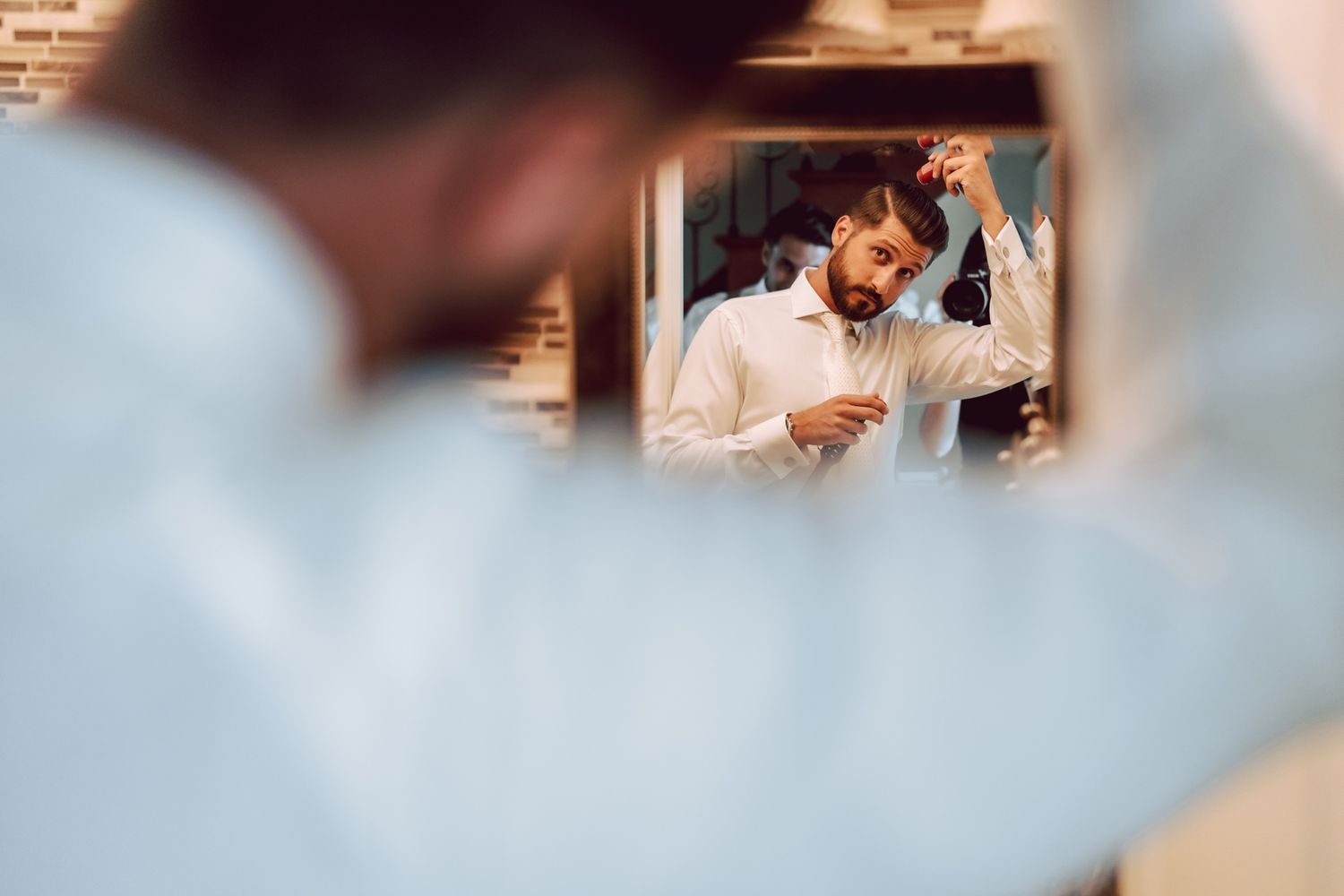 Bearded man in white shirt adjusting hair while looking in mirror, reflected in elegant setting.