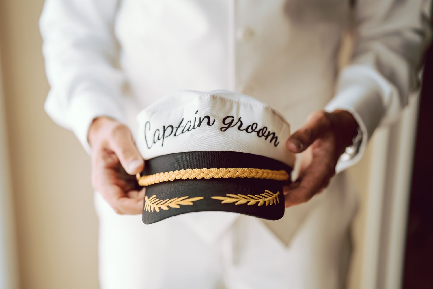 Groom holding a white captain's hat with gold trim reading Captain Groom on his wedding day.