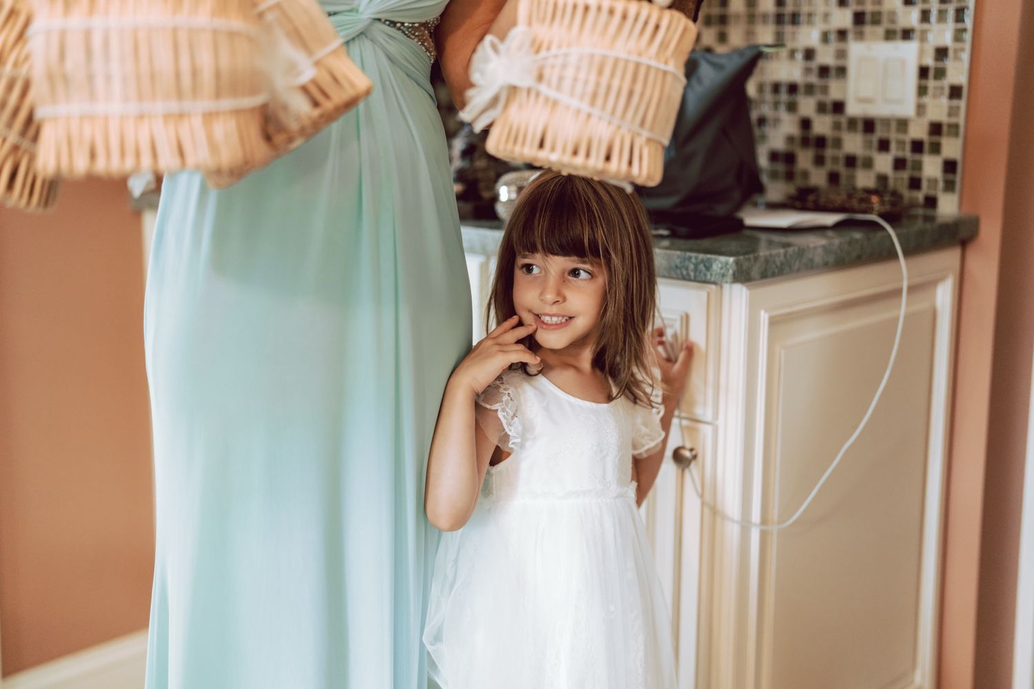 A young flower girl in a white dress smiles shyly beside a woman in a mint gown during wedding preparations.