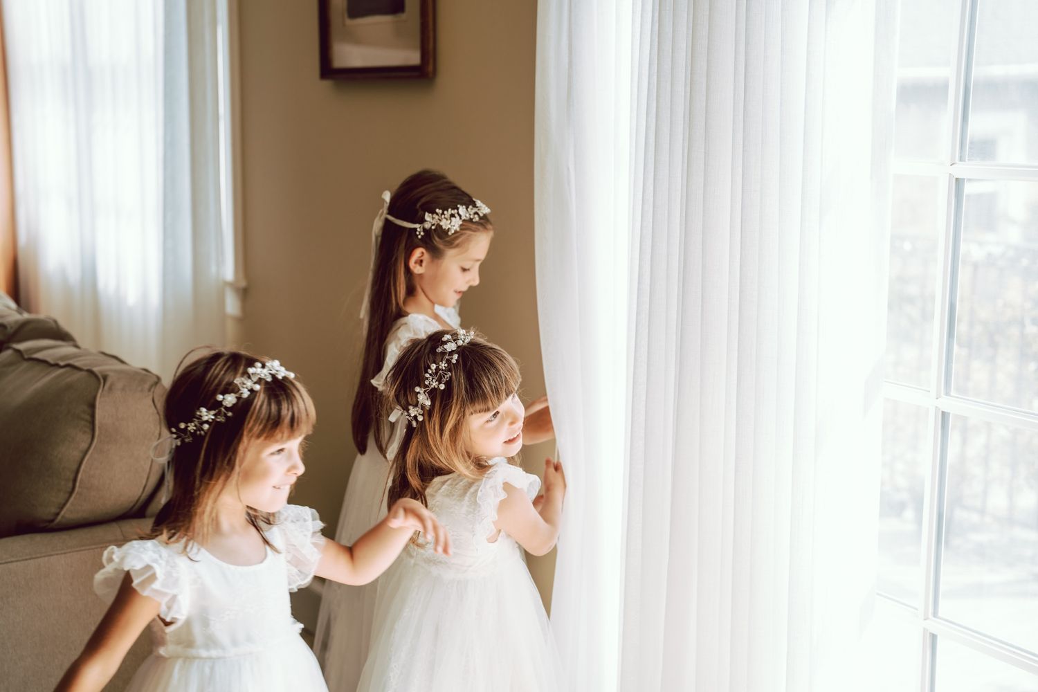 Three young flower girls in white dresses and floral hair wreaths stand by a sunlit window.