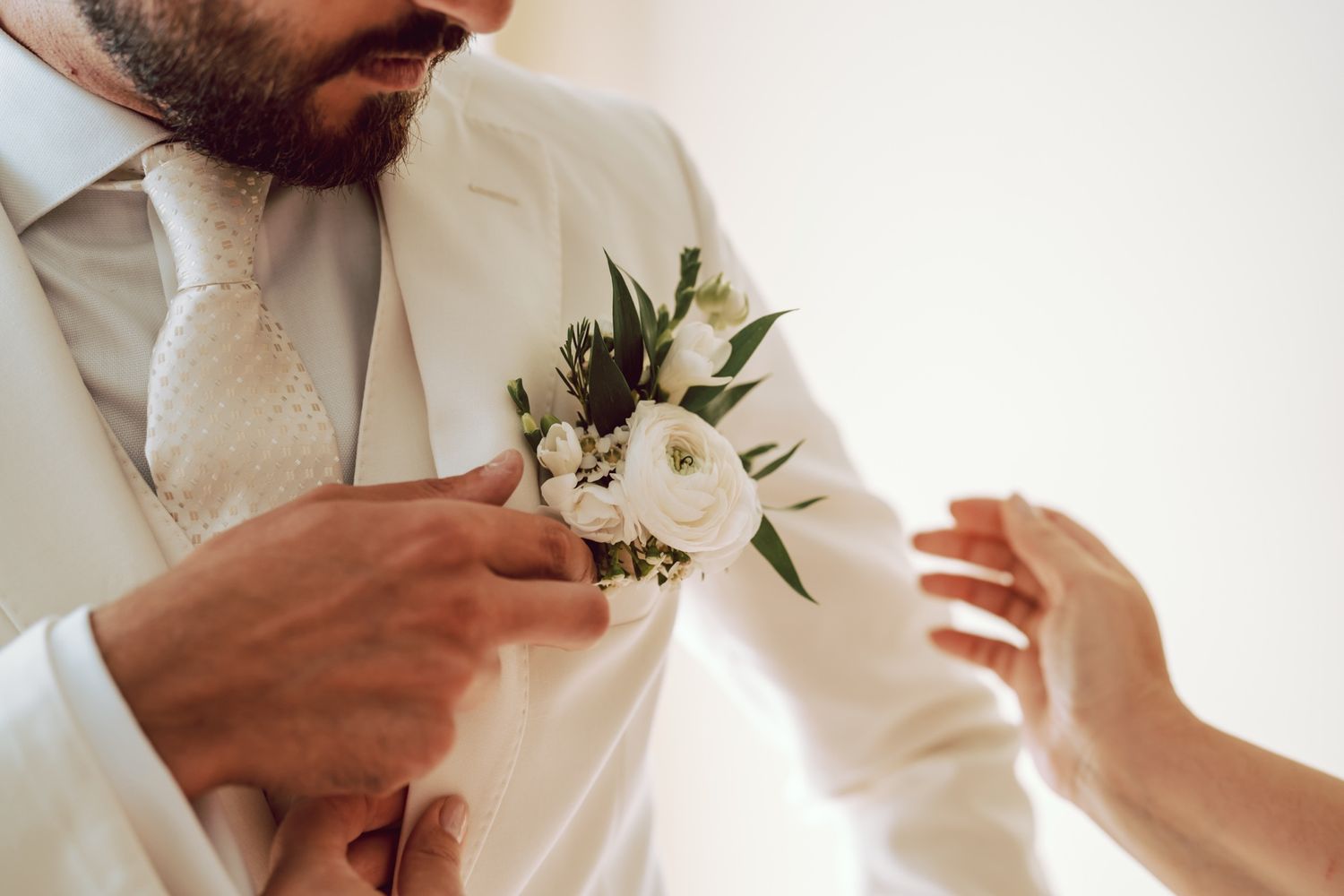 Groom in white suit having boutonniere with white flowers pinned to lapel on wedding day.