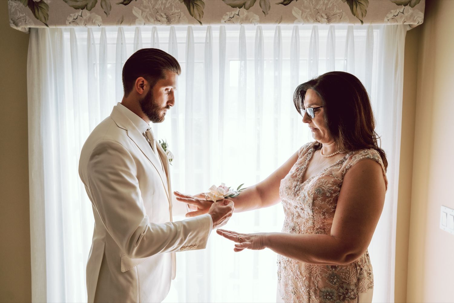 Groom in white suit receiving wedding rings from woman in lace dress by window before ceremony.