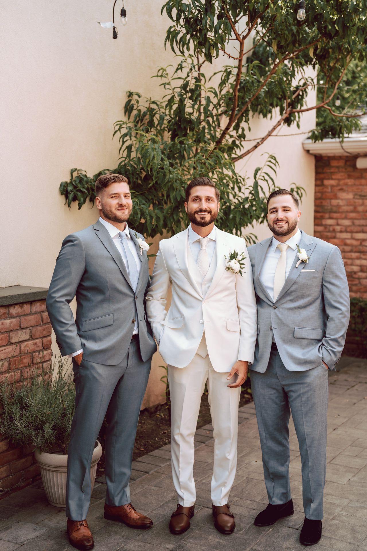 Groom in white suit poses with two groomsmen in light gray suits and boutonnieres outdoors at a wedding.