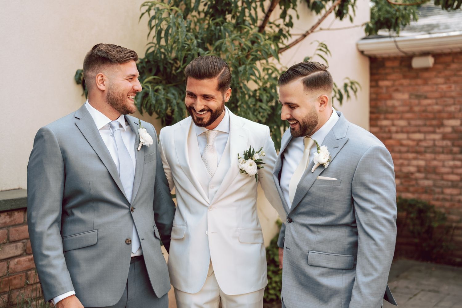 Groom in white suit stands with two groomsmen in matching gray suits and boutonnieres, laughing outdoors.