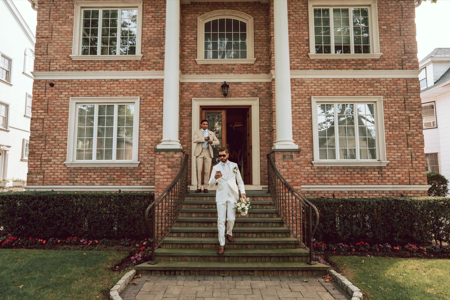 Groom in white suit walks down steps of elegant brick mansion holding bouquet on wedding day.