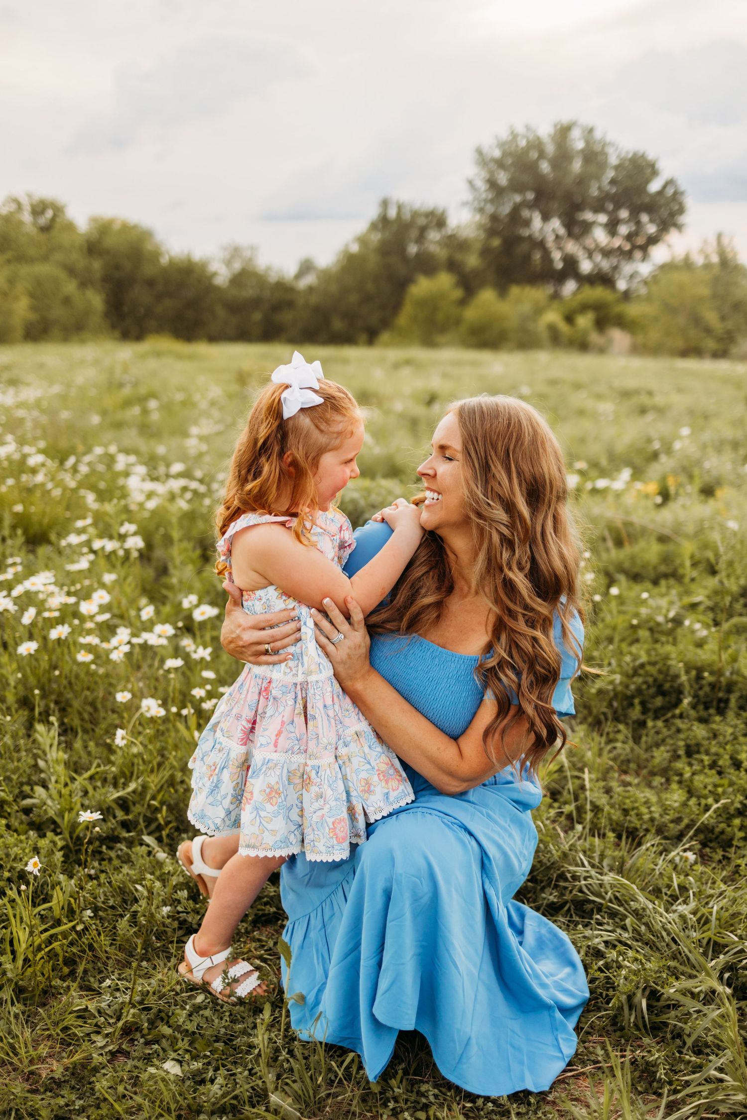 A smiling mother in a blue dress kneels in a flower field, lovingly holding her young daughter in a floral dress.