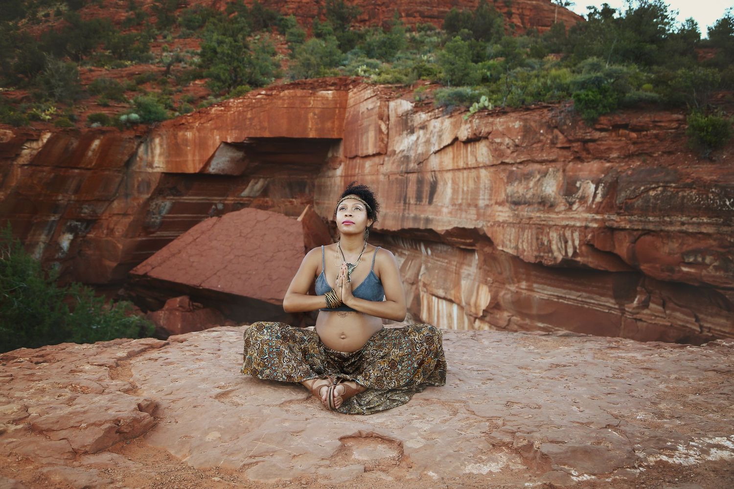 Pregnant woman sits cross-legged in a meditation pose on red sandstone rock surrounded by Sedona-style canyon formations.