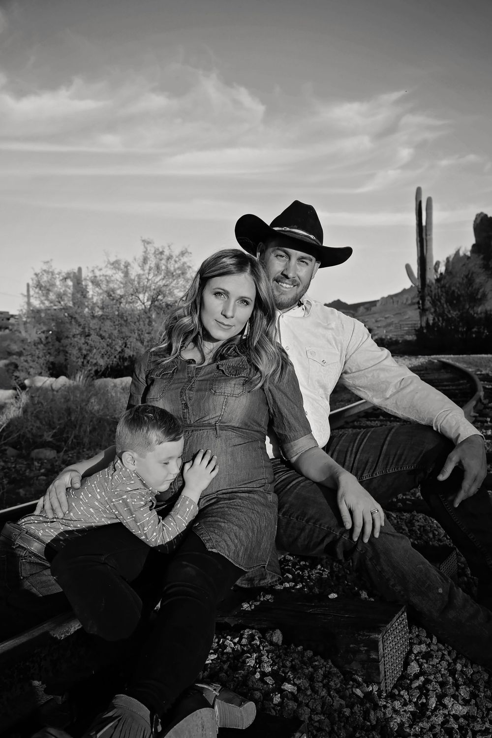 Black and white family portrait of a cowboy-hatted man, pregnant woman and young boy sitting among desert cacti.