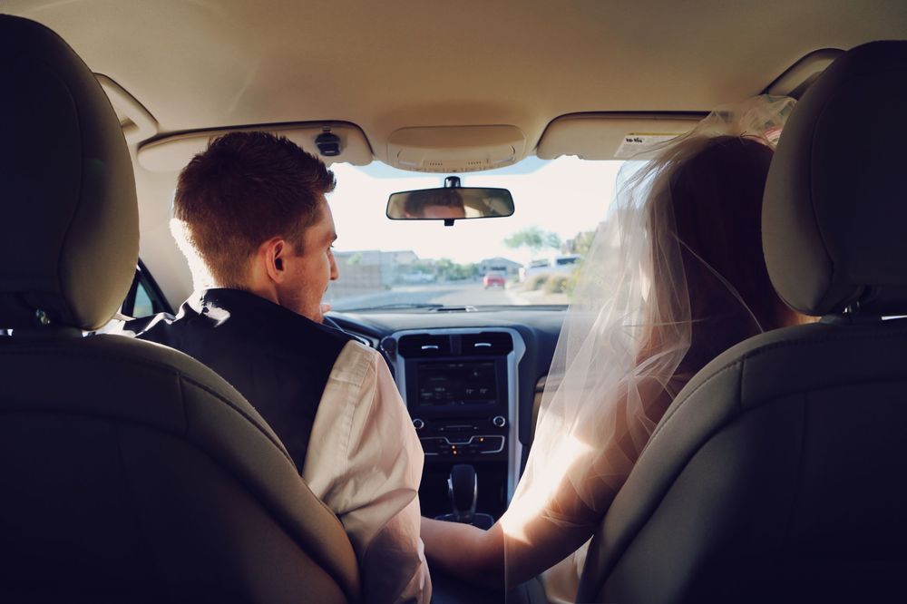 A groom drives a car while the bride in a veil sits beside him, seen from the back seat on a sunny day.