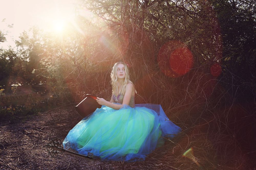 A young woman in a teal and blue tulle gown sits holding a book in a sunlit forest with colorful lens flares.