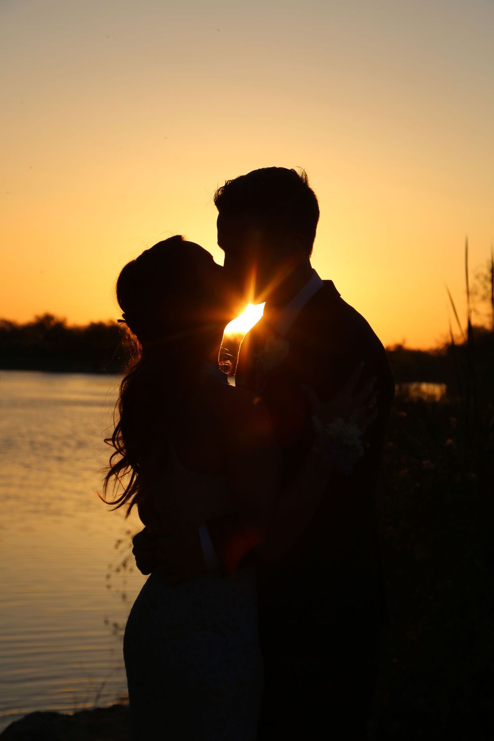 Silhouette of a couple kissing by a calm lake as the sun sets in a glowing orange sky.