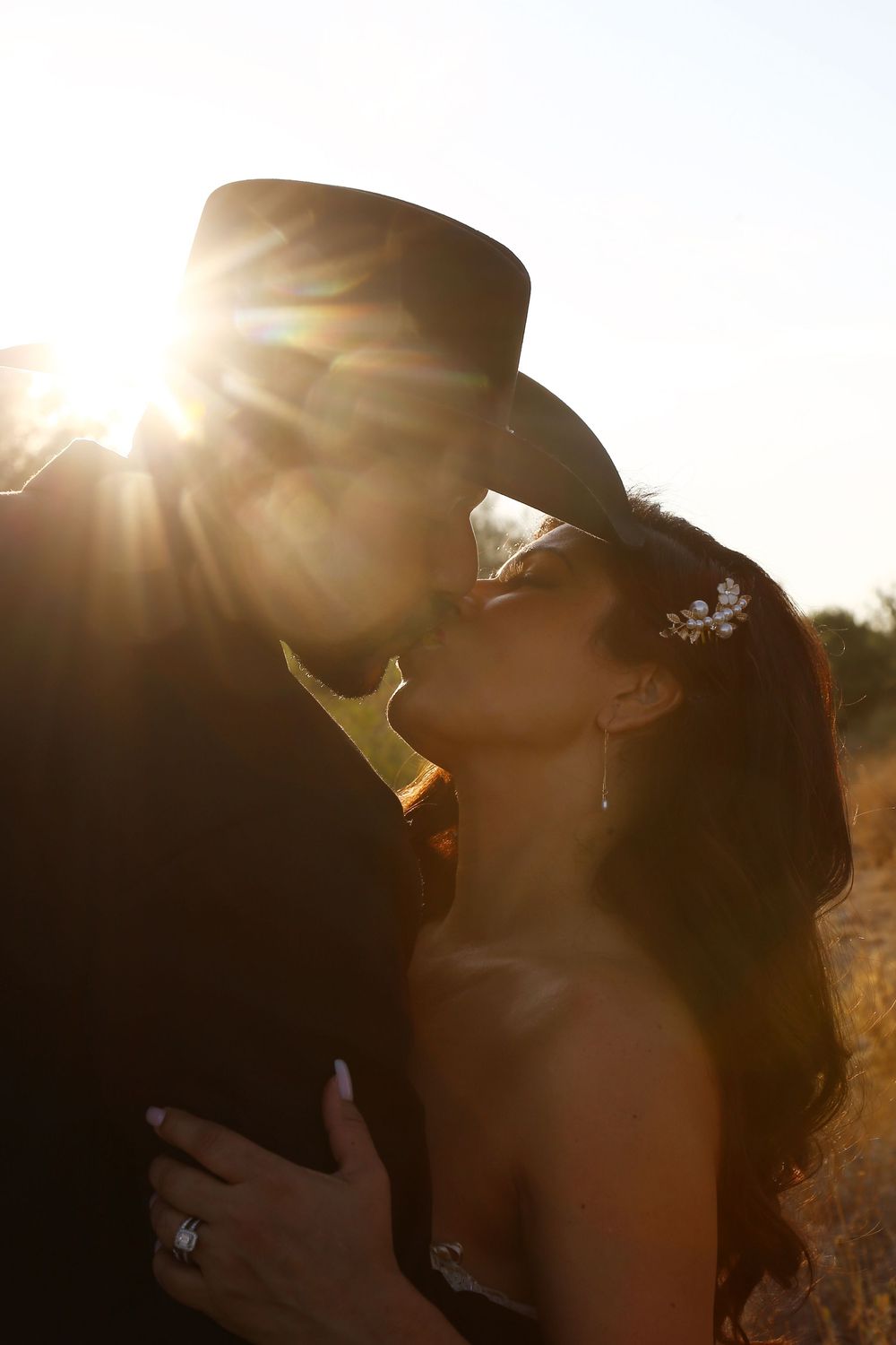 A cowboy-hatted groom kisses his bride, who wears a floral hair piece, in golden backlit sunshine.
