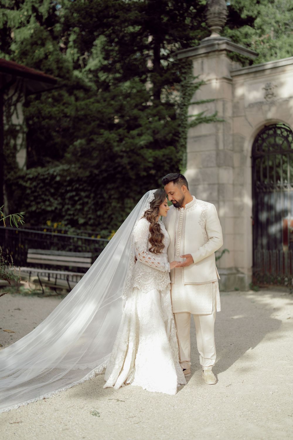 Bride and groom in elegant white wedding attire stand together outdoors near ornate iron gates surrounded by lush greenery.