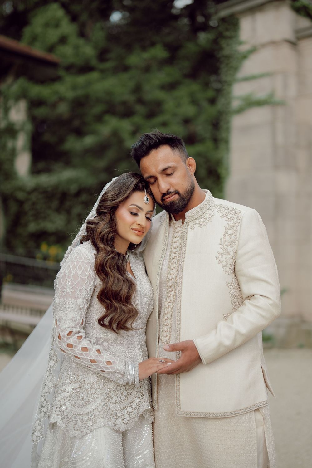 South Asian bride and groom in elegant ivory wedding attire share a tender romantic moment outdoors.