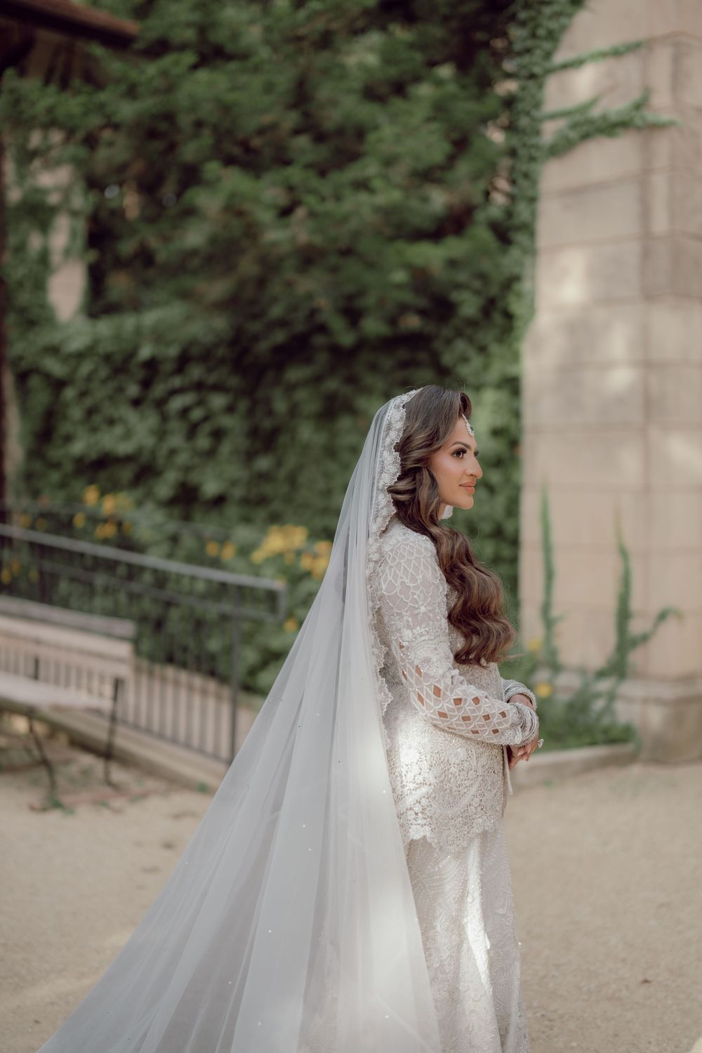 Elegant bride in a beaded long-sleeve wedding gown and cathedral veil poses in a lush garden setting.