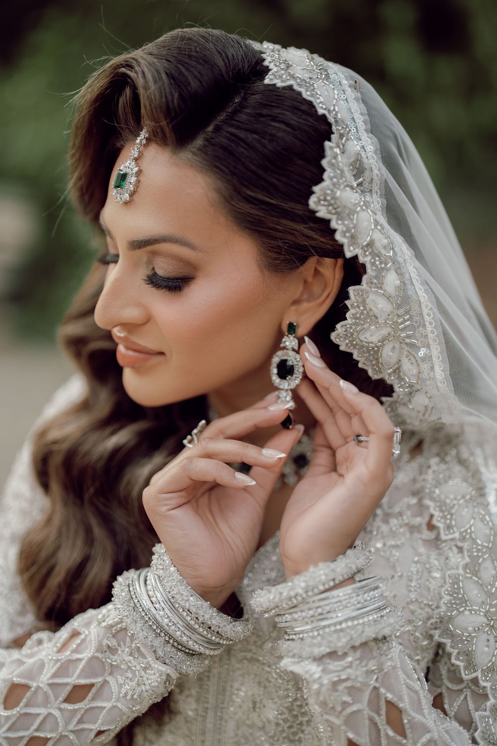 A bride in white embroidered lehenga adjusts her earring, wearing a maang tikka and lace dupatta.
