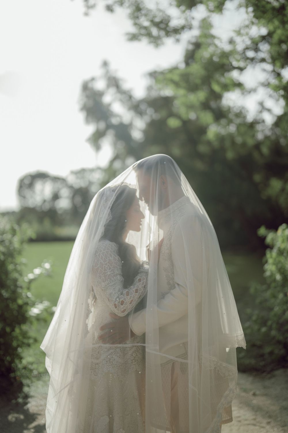 A bride and groom share an intimate moment under a flowing white veil outdoors, surrounded by lush green trees.