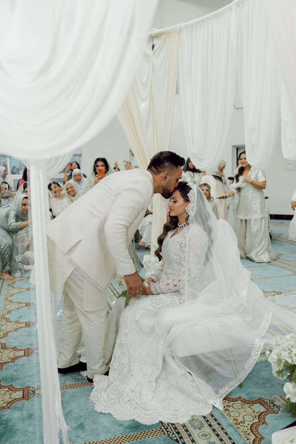 Groom in white suit kisses bride in lace wedding dress under draped canopy at elegant Malay wedding ceremony.