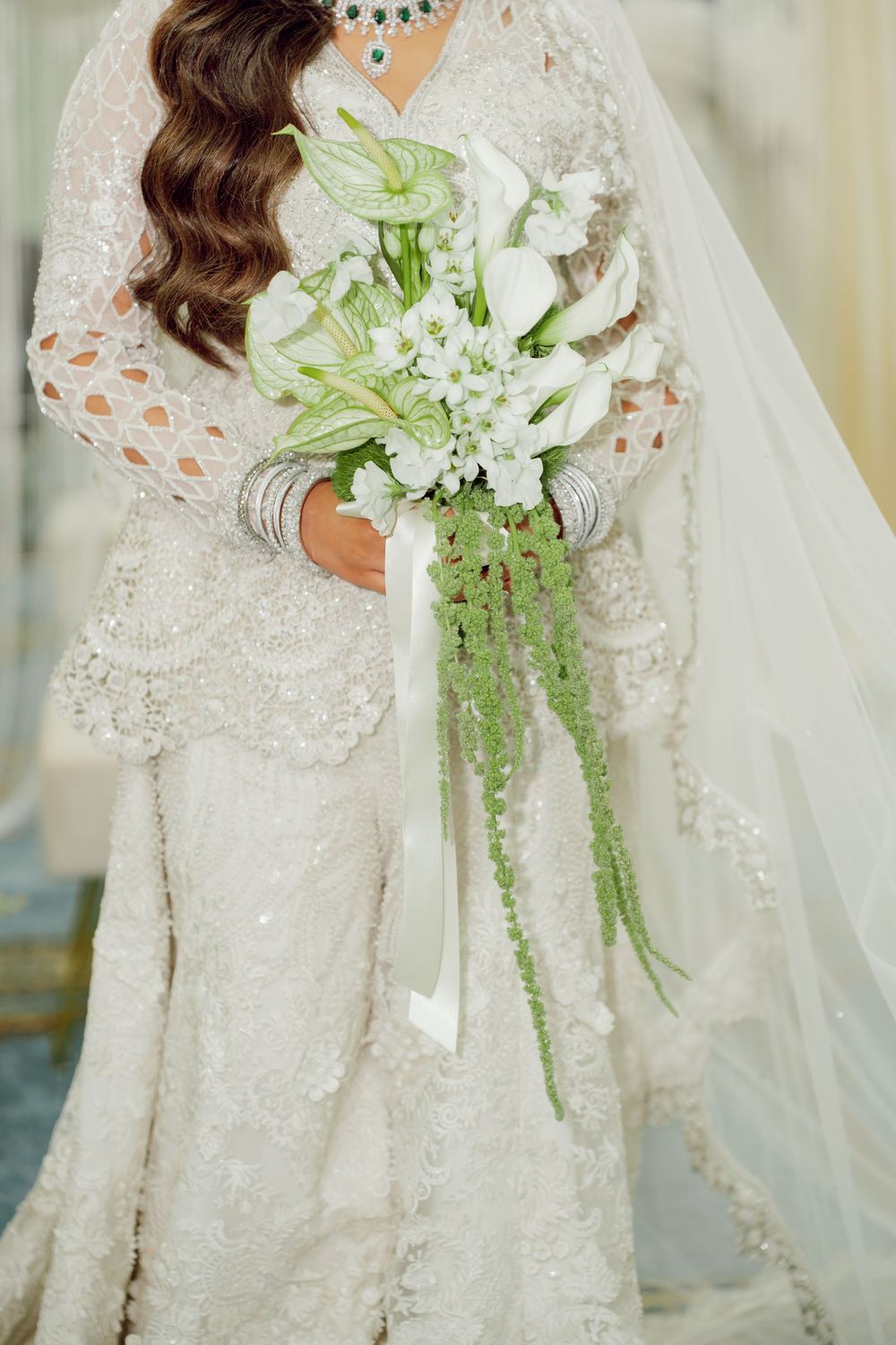 Bride in beaded lace gown holding white calla lily bouquet with green trailing accents and ribbon.