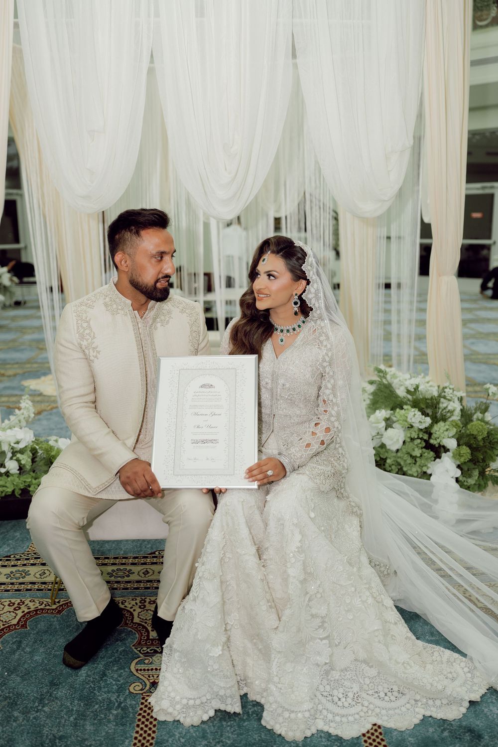 Elegant couple in white wedding attire holding marriage certificate at decorated ceremony venue with floral arrangements.