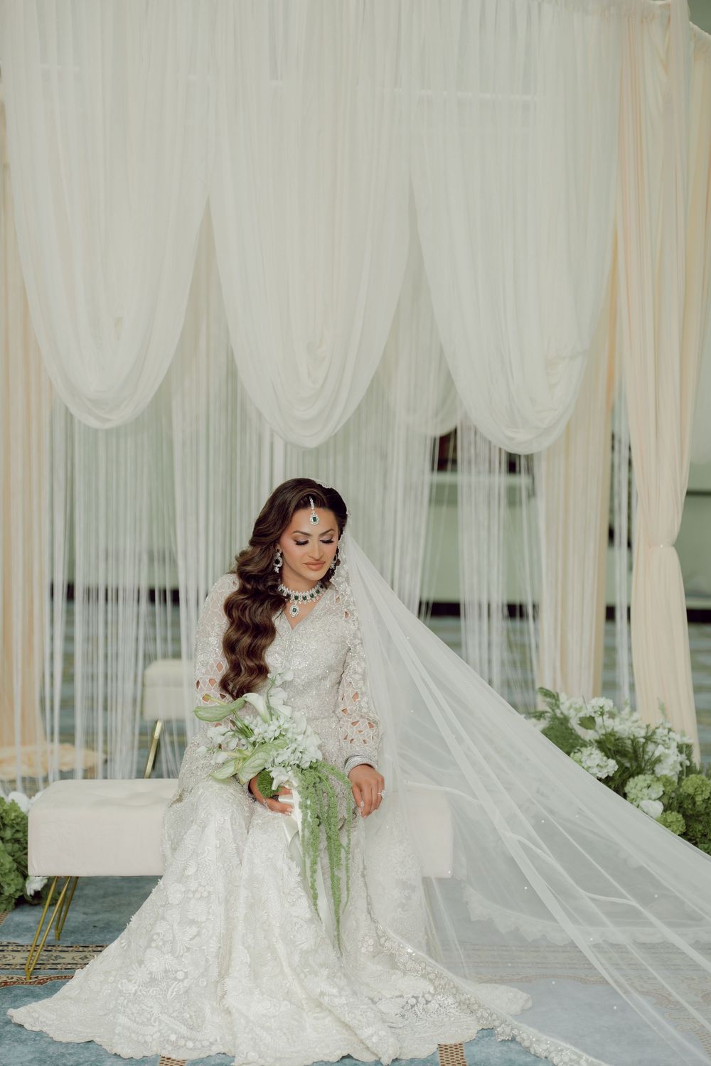 Bride in white lace gown with long veil sits elegantly before draped fabric backdrop holding cascading bouquet.