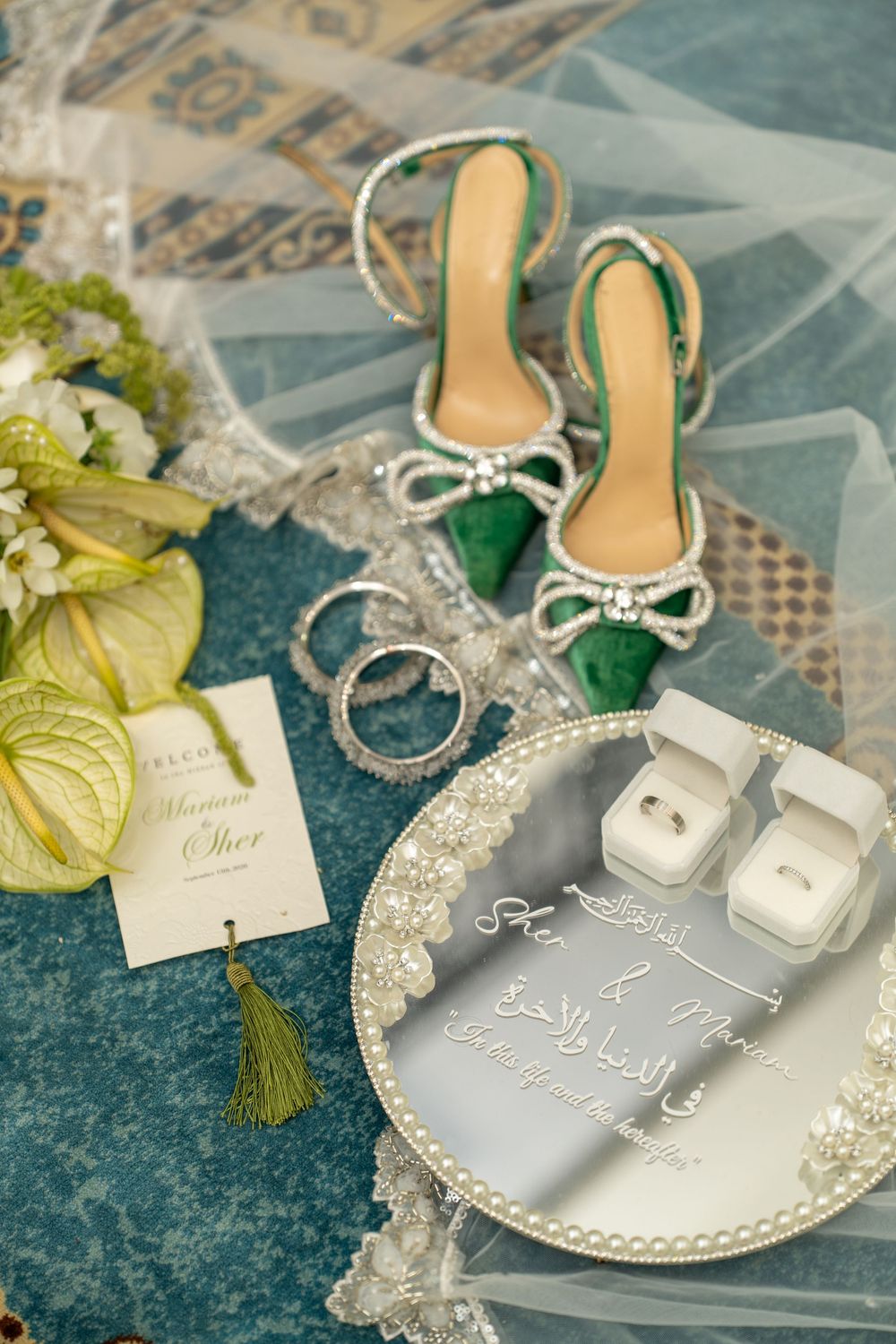 Elegant wedding flat lay with green heels, diamond rings, pearl tray, bouquet, and Arabic calligraphy invitation on teal surface.