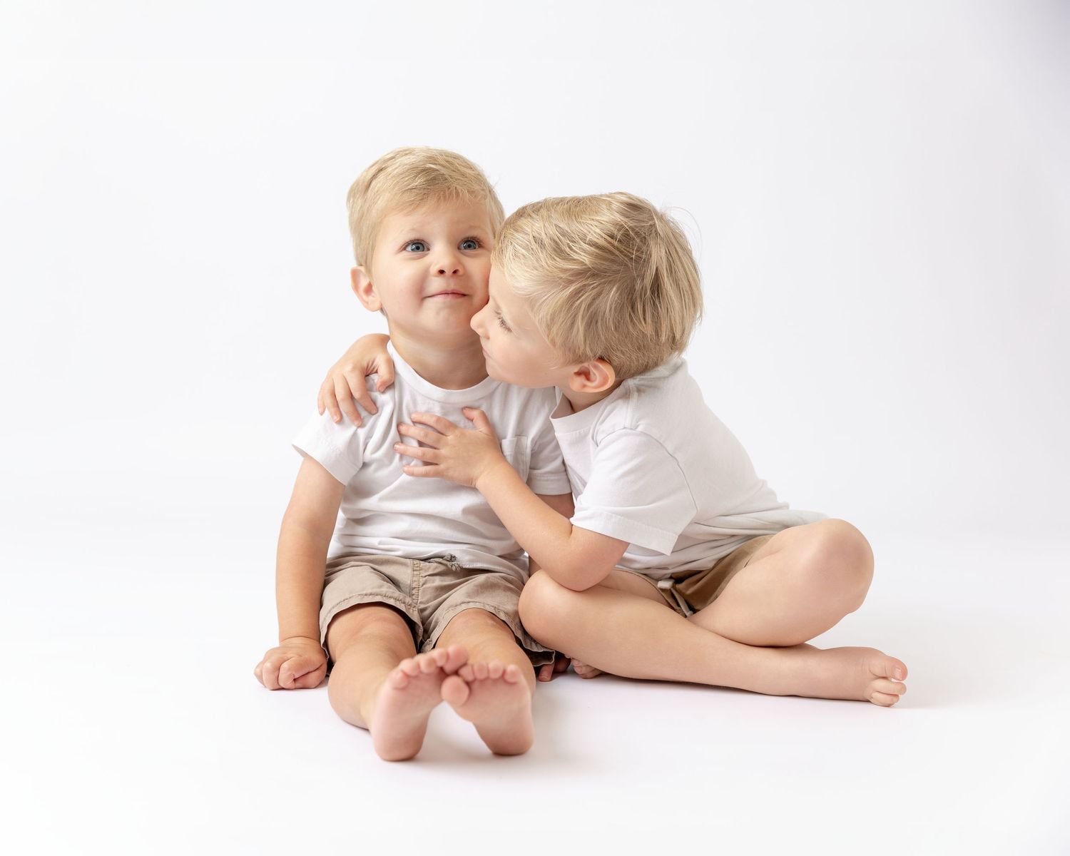Two young blonde boys in white shirts, older child kissing younger sibling on cheek while sitting together on white background.